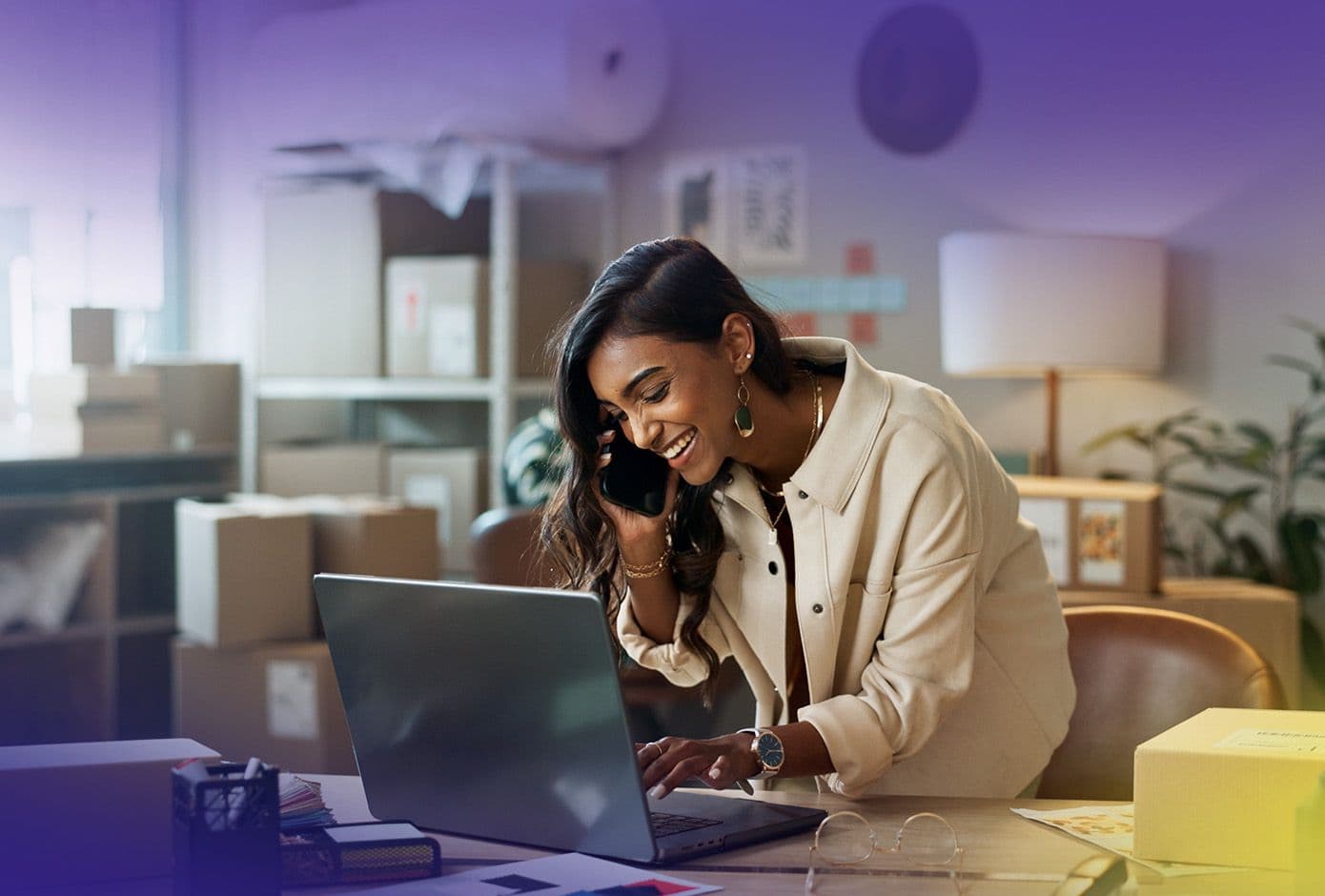 Woman on phone while typing on laptop, leaning over desk with lots of boxes and shipping materials behind her.