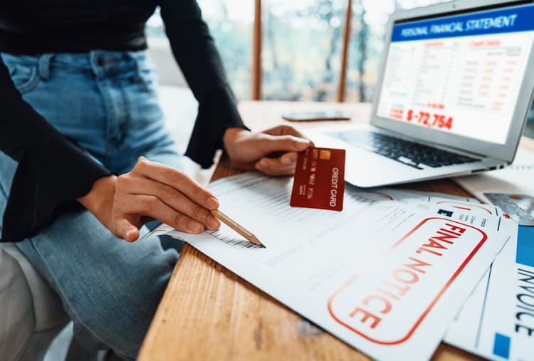 Person holding a credit card while looking at an overdrawn bank statement on a laptop screen and the final notice of a paper bill.