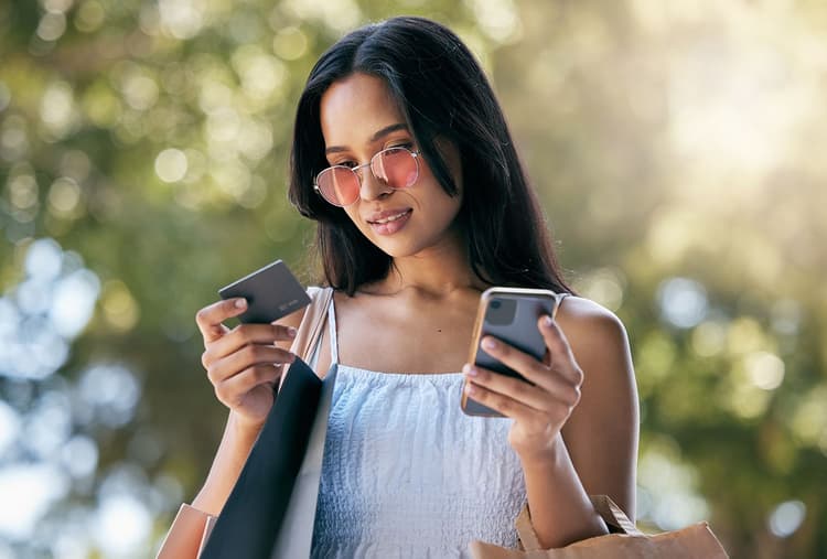 Woman dressed in a tank top and sunglasses looks at her credit card and phone while carrying shopping bags.