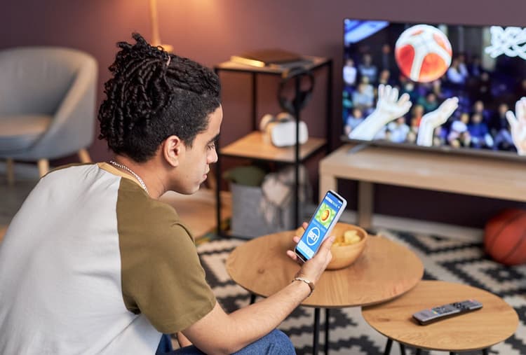 Gen Zer places bet on his mobile phone while a basketball game plays on the tv.